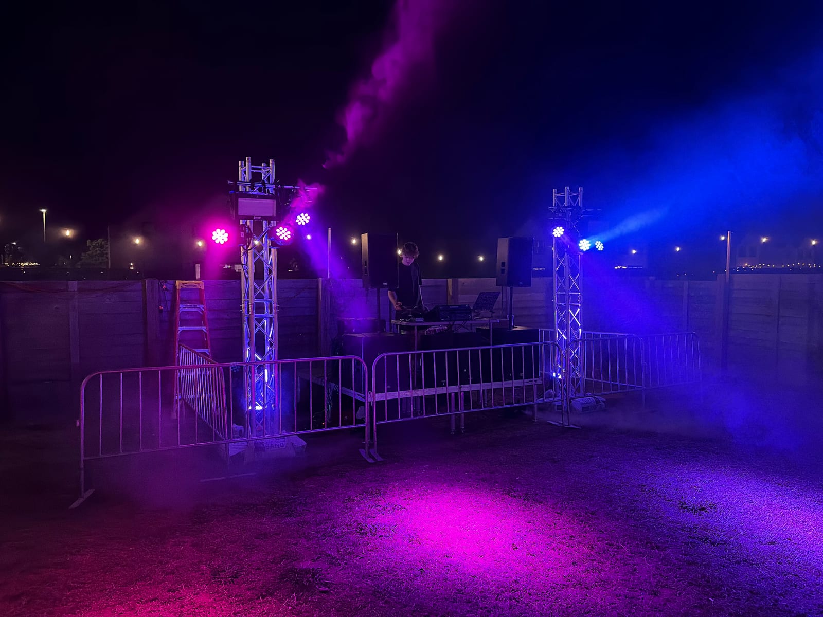 Truss towers throwing magenta and blue light through haze over an outdoor DJ stage at night